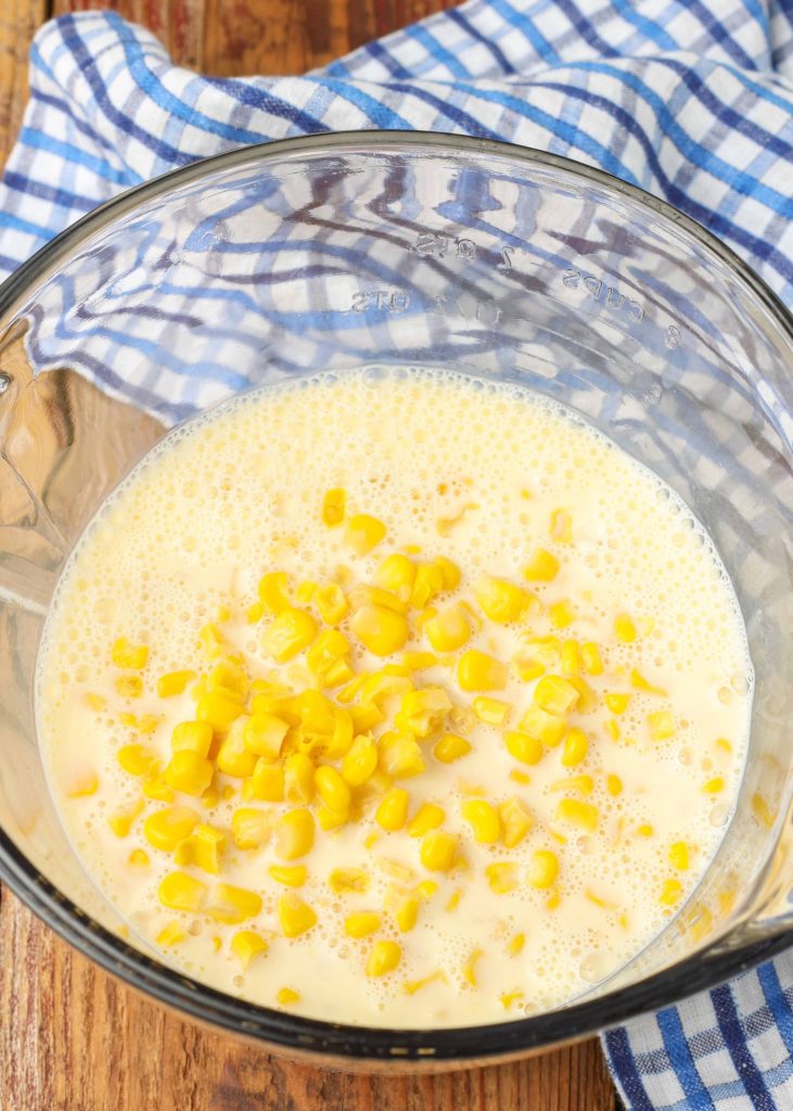 Milk and corn have been added to a clear glass mixing bowl, set atop a blue and white checkered tea towel.