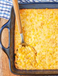 a long handled metal serving spoon has been inserted into the scalloped corn in a metal casserole baking pan.