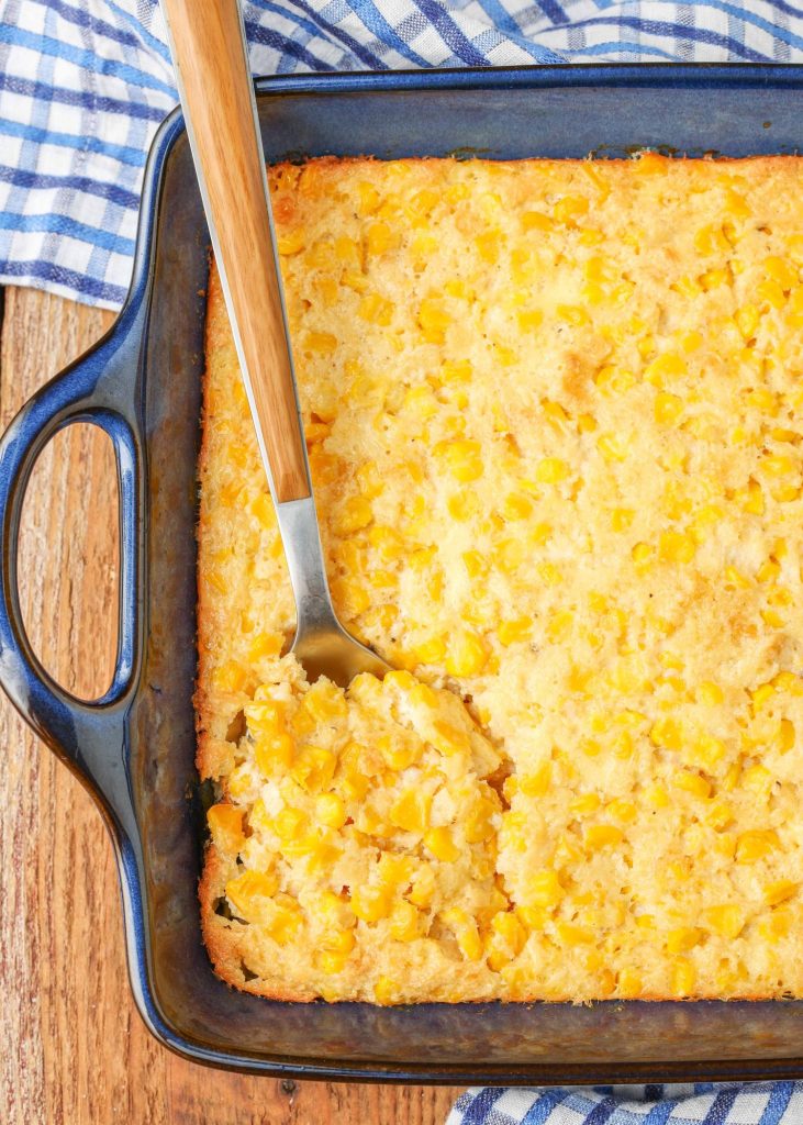 a long handled metal serving spoon has been inserted into the scalloped corn in a metal casserole baking pan.
