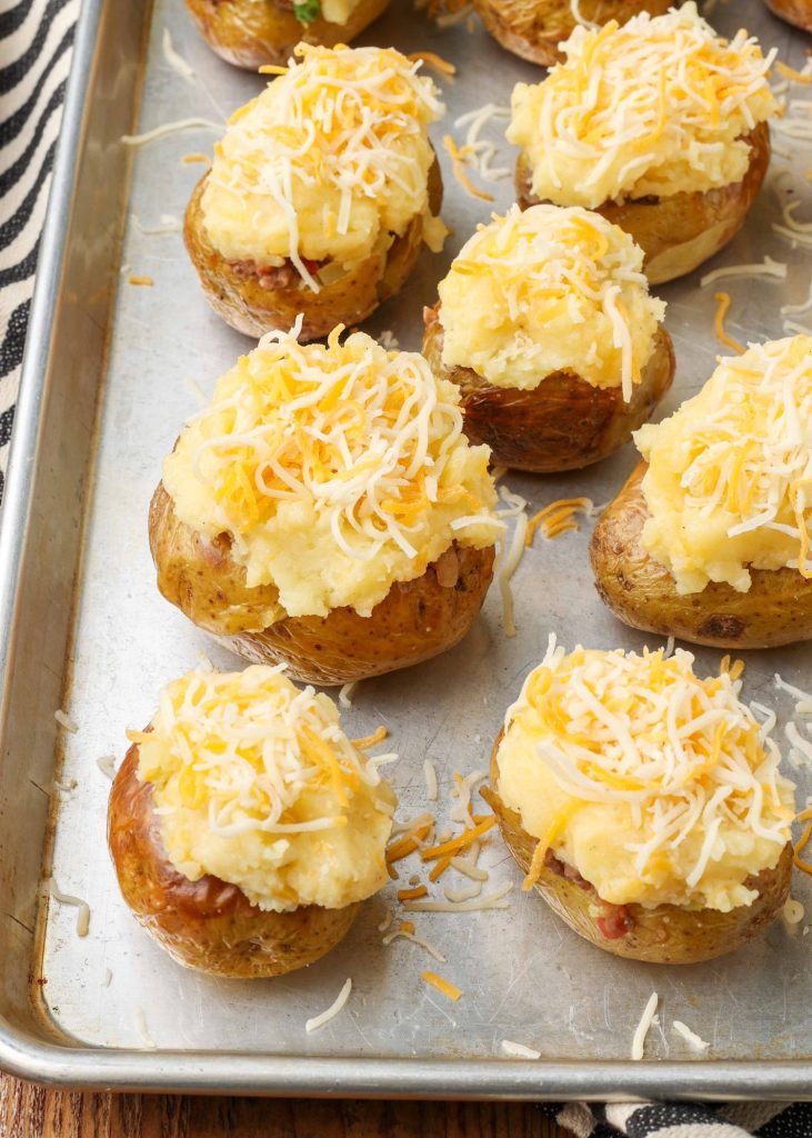 All of the baked potatoes have been topped with mashed potatoes on the metal sheet pan, and sprinkled with cheese, ready to go back into the oven.