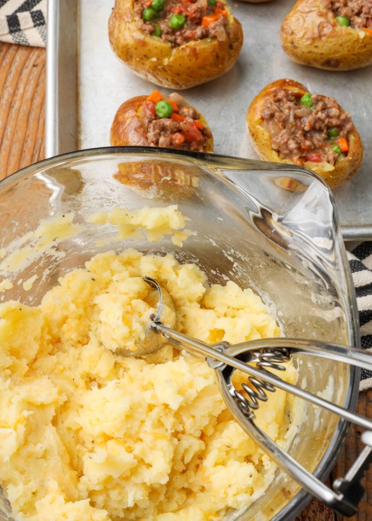 The mashed potato topping is in a clear glass bowl beside the sheet pan loaded with potatoes.