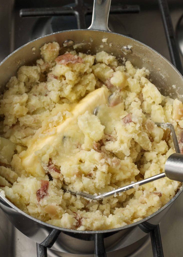 Overhead vertical shot of potatoes mid-mashing and butter, cooking in a sauce pan with a silver potato masher