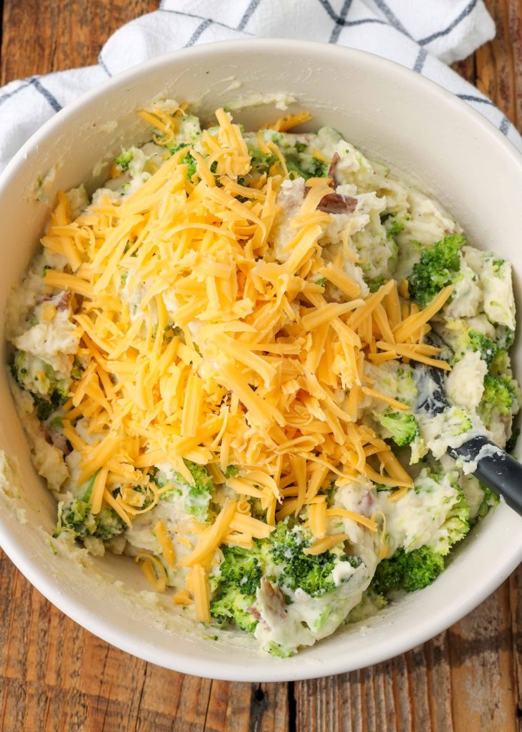 Overhead shot of broccoli mashed potatoes mixture topped with shredded cheese, served in a white bowl with a striped white and black hand towel