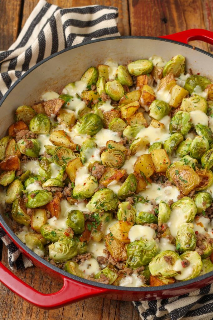 Overhead zoomed-out brussels sprouts with sausage and potatoes, served in a red dish with a striped black and white hand towel