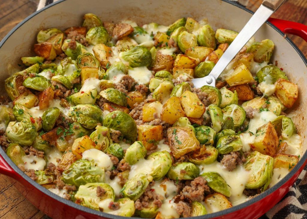 Brussels sprouts with sausage and potatoes, served in a red dish with a silver spoon