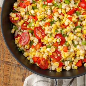 A top down photo of a bowl filled with fresh corn salad, with corn kernels, grape tomatoes, red onions, and bell peppers visible.