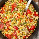 A vertically aligned photo of a serving bowl of corn salad with a long-handled metal spoon.