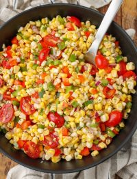 A vertically aligned photo of a serving bowl of corn salad with a long-handled metal spoon.