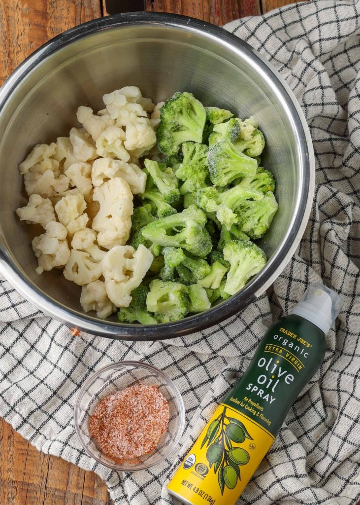 frozen broccoli and cauliflower in metal bowl with olive oil spray and spices in smaller bowl