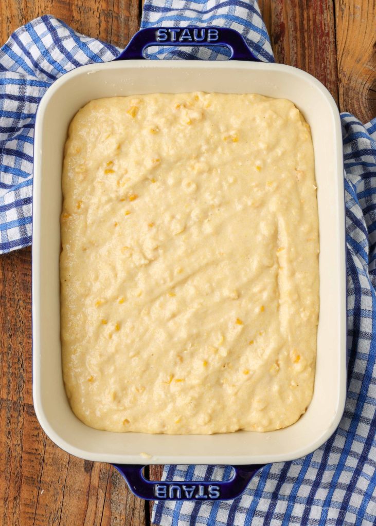 A baking dish with a white inner and royal blue outer colors, is filled with corn spoon bread mixture, ready to go into the oven.