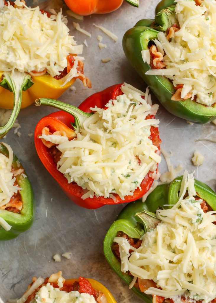 Stuffed peppers with shredded cheese on top, ready for the oven, are visible on a metal baking sheet in this top down photo.
