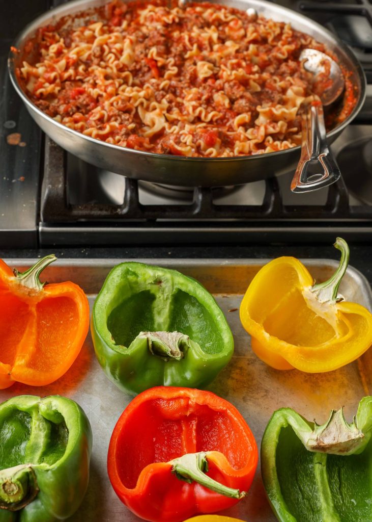 Bell pepper halves on a metal baking sheet are in the foreground with meat sauce and pasta visible in the background in a pan.