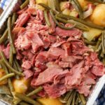 A vertically aligned photo of a white baking dish filled with chunks of ham, potatoes, and green beans over a blue and white checkered tea towel. A horizontal close up photo of chunks of ham, green beans and yellow potatoes. A vertically aligned close up photo of chunks of ham, potatoes and green beans with just the edge of a blue and white checkered tea towel visible in the top left and bottom right corner. A black oval crock pot top down image vertically aligned. The Crock pot is loaded with