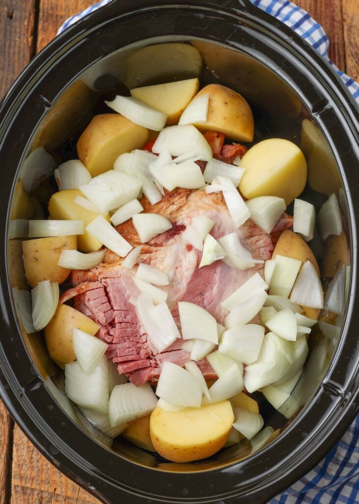 The ham is just visible beneath the chunks of onion and potatoes in the crock pot.