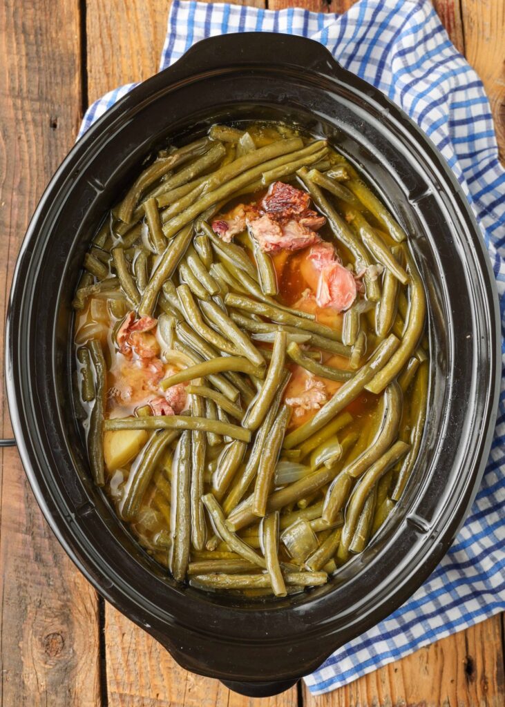 A top down photo of a black oval crockpot loaded with green beans, ham, and potatoes, covered with chicken stock. Ready to cook!