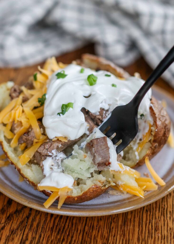A black metal fork has a bite sized portion of the loaded steak baked potato.