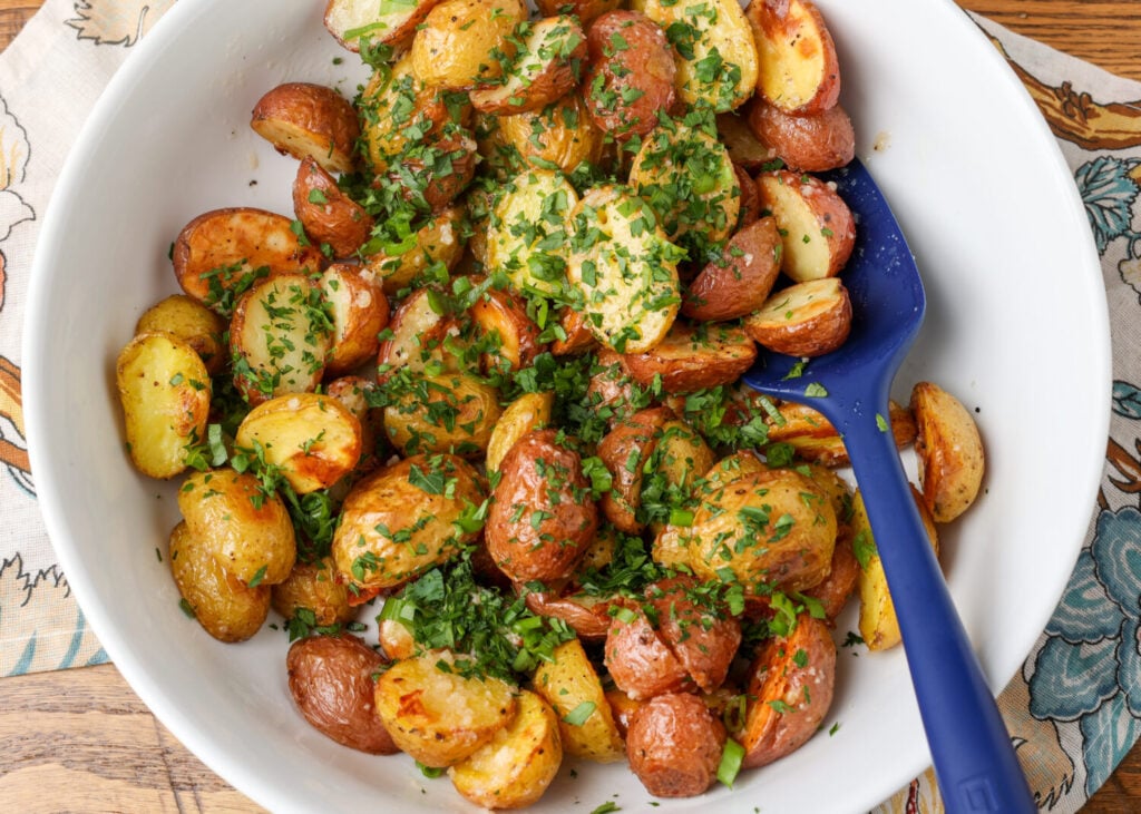 A close up image of roasted potatoes in a white bowl sprinkled with fresh herbs.