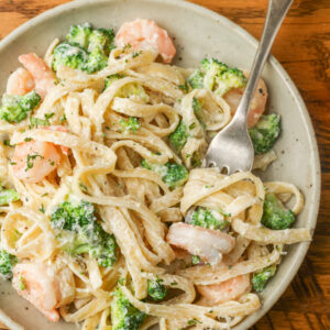 A metal fork has twirled a bite of pasta from the ceramic plate full of shrimp and broccoli pasta. The fork handle proceeds from the center of the image toward the top right corner. A wooden tabletop is visible beneath the plate.