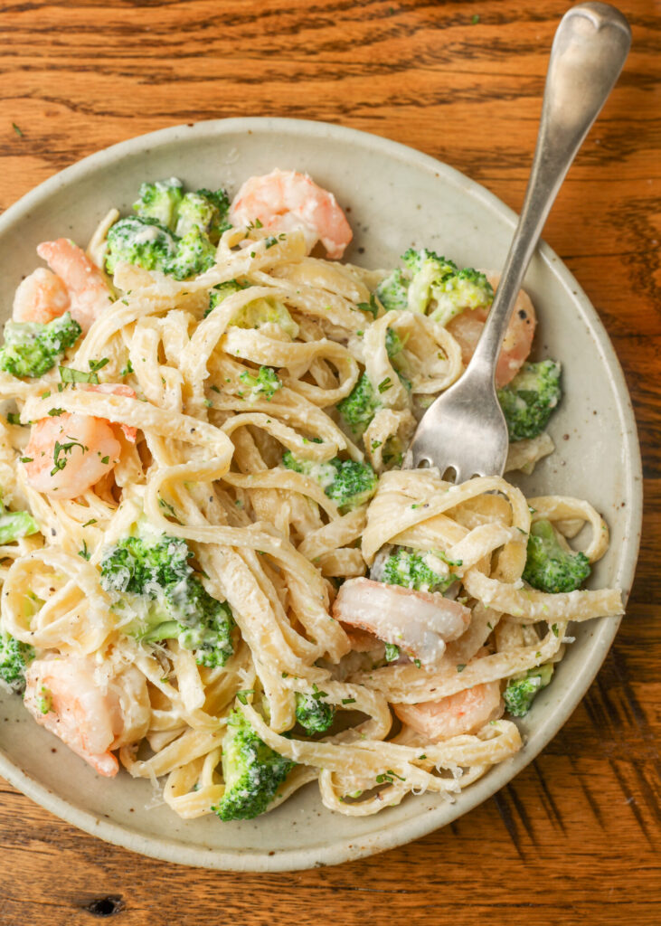 A metal fork has twirled a bite of pasta from the ceramic plate full of shrimp and broccoli pasta. The fork handle proceeds from the center of the image toward the top right corner. A wooden tabletop is visible beneath the plate.