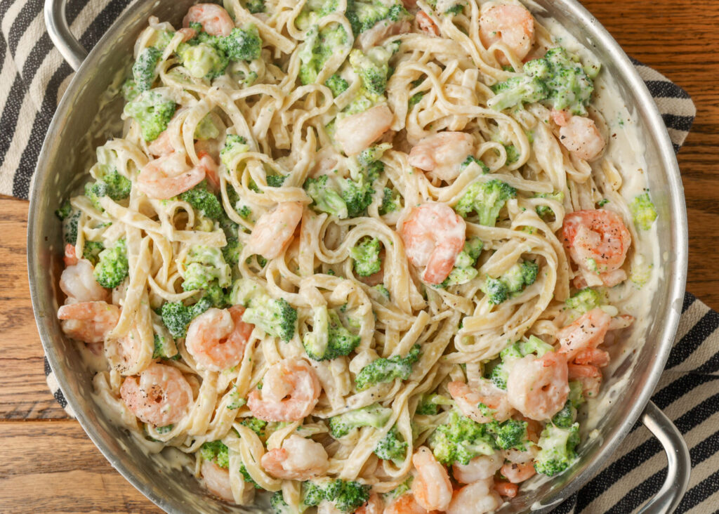 A close up, top down, horizontal image of a metal pan filled with creamy shrimp and broccoli pasta, ready to be served.