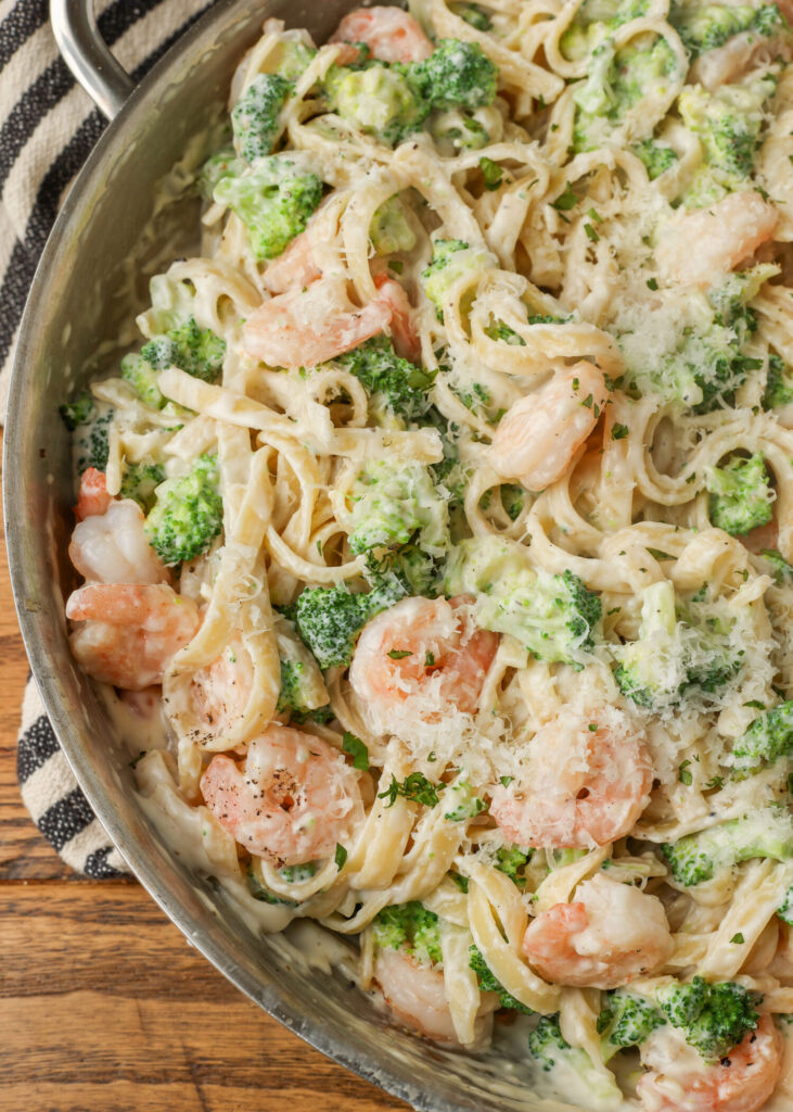 A close up vertical image of the left half of a metal pan, full of delicious pasta with shrimp. A black and white striped towel is visible in the background.
