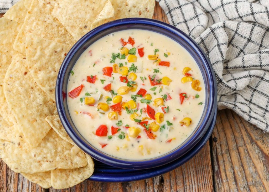 queso dip with corn and bell peppers in blue bowl with tortilla chips and plaid towel