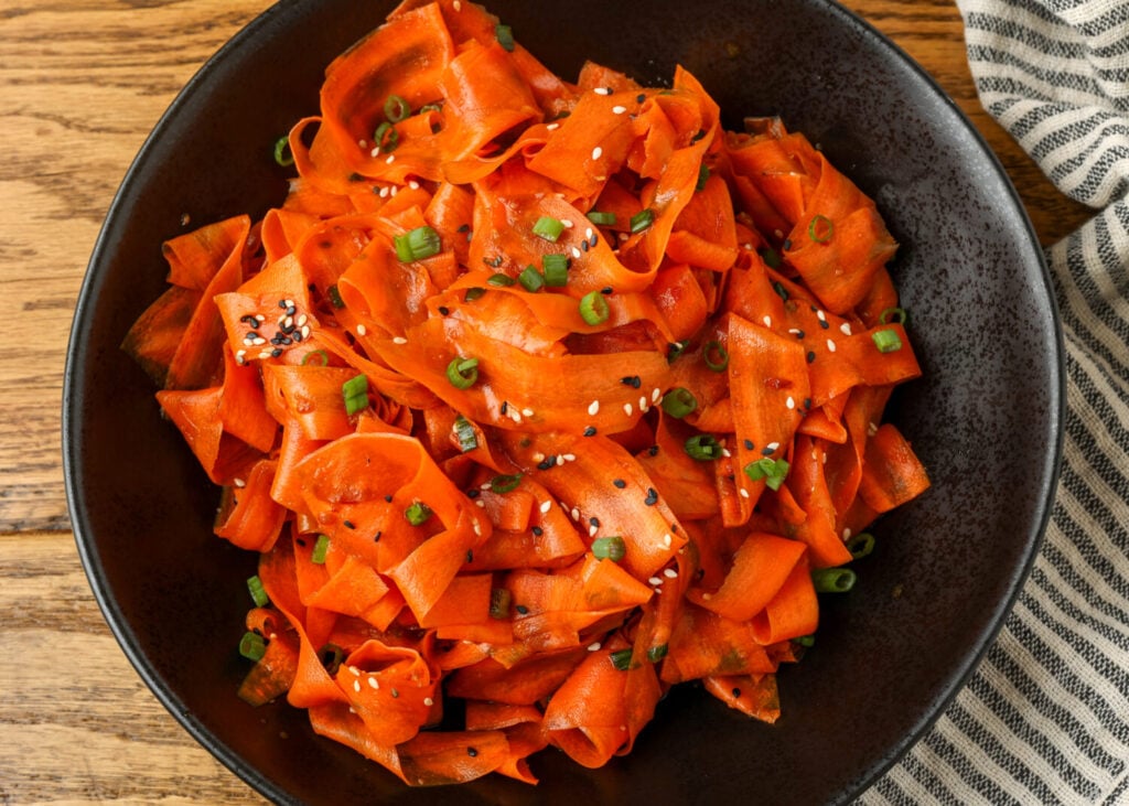 Vivid orange carrots fill a black ceramic bowl over a wooden tabletop in this Asian Carrot Salad.
