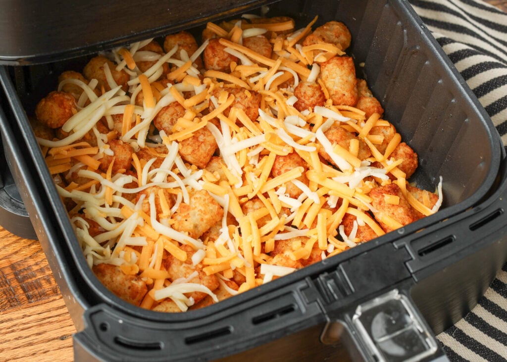 Freshly shredded cheddar cheese scattered over crispy air fryer tater tots in the basket before the final melt.