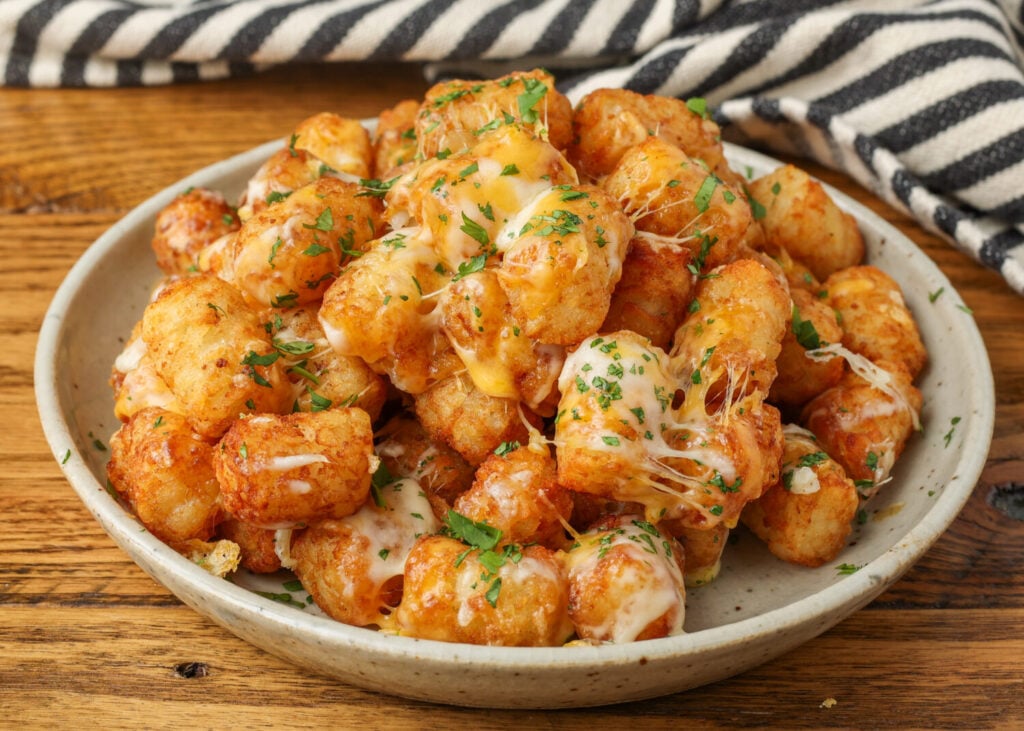 A pile of crispy air fryer tater tots sits on a ceramic beige plate, sprinkled with fresh herbs and melted cheese.