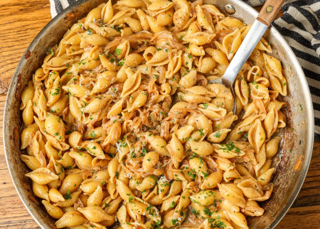 A top down image of a pan with creamy sauce and pasta and a wooden handled long metal spoon.