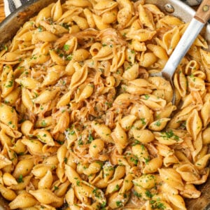 A top down image of a pan with creamy sauce and pasta and a wooden handled long metal spoon.