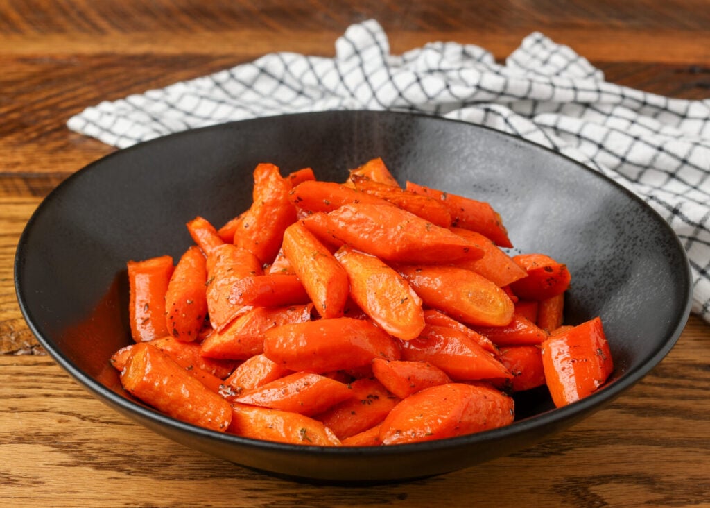 Vivid orange carrots, glazed with a honey sauce are piled high in a ceramic black bowl over a wooden tabletop.